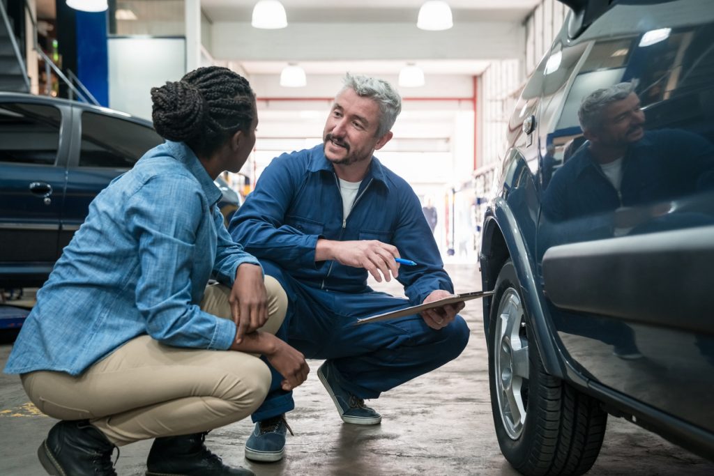 Car mechanic at a successfully built body shop inspecting vehicle with customer checking car wheel in the auto repair shop, holding clipboard and talking with customer